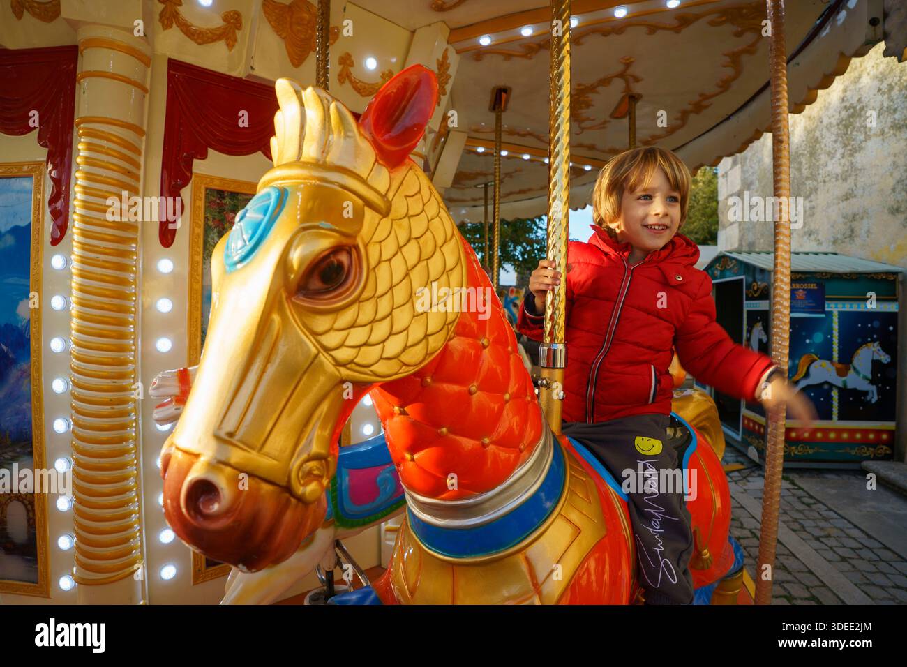 Enfant souriant en veste rouge chevauchant un cheval de carrousel coloré dans un parc d'attractions en plein air pendant la journée Banque D'Images