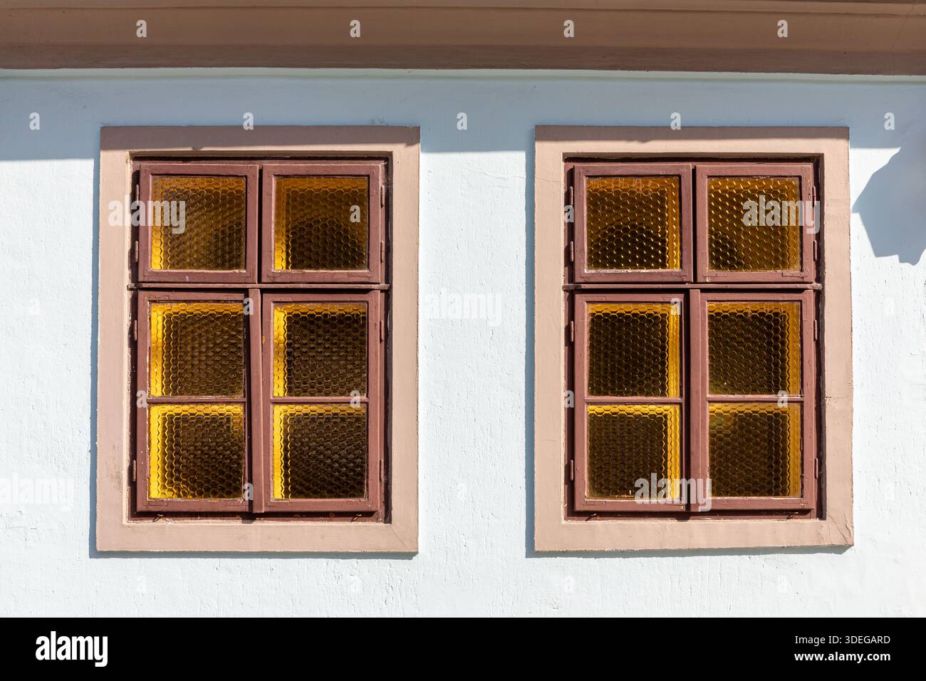 Deux fenêtres de style vintage avec des cadres en bois et des panneaux de verre texturés de couleur ambre divisés en quatre sections, sur une façade blanche avec cadre brun. Banque D'Images