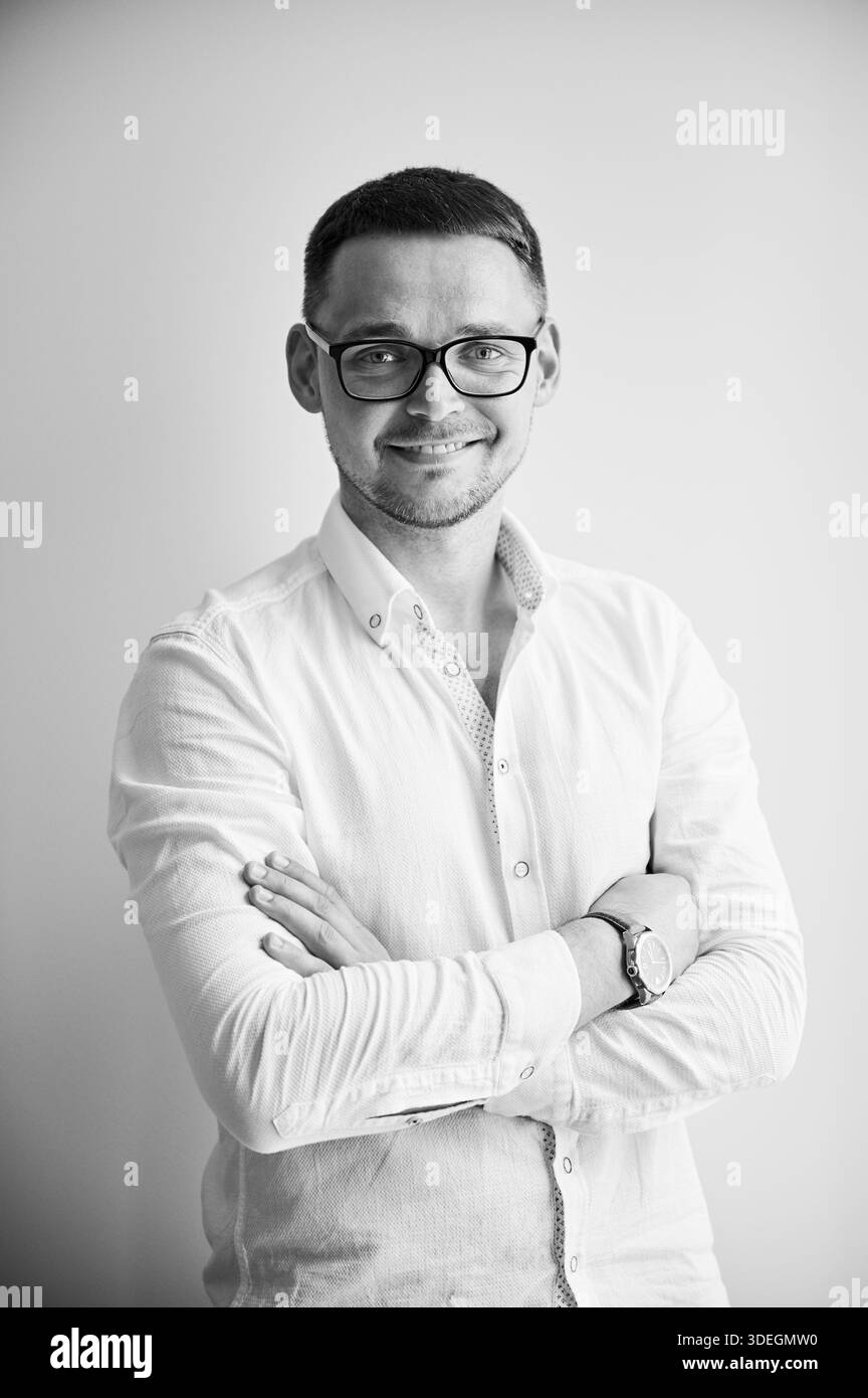 Charmant homme souriant directement à la caméra. Portrait de beau jeune homme dans des vêtements décontractés gardant les bras croisés tout en se tenant debout sur fond blanc. Image en noir et blanc. Banque D'Images