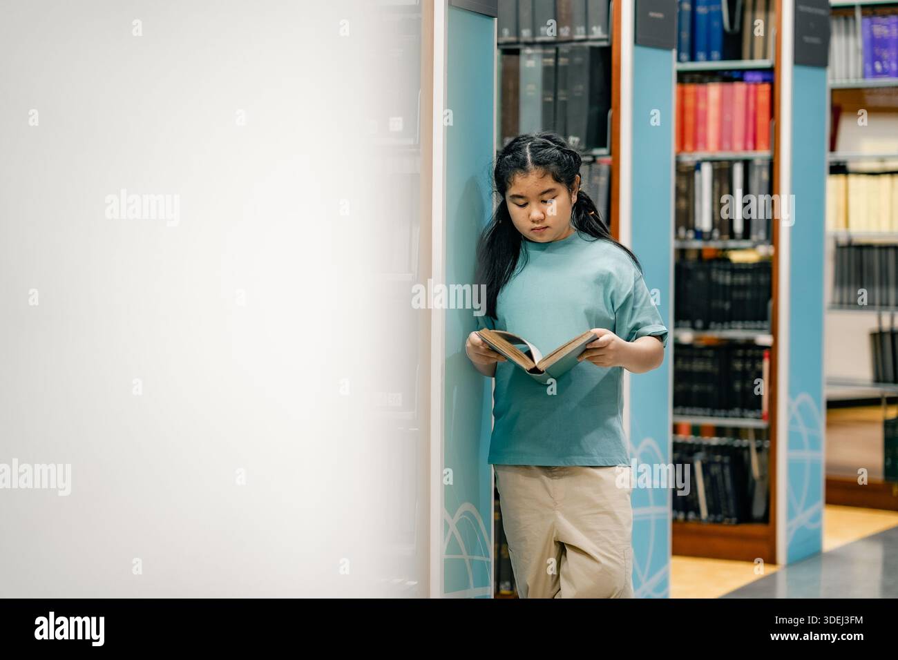 Enfant retournant des pages de livre à l'intérieur de l'allée de la bibliothèque de l'école. Posture calme communique concentration auto-apprentissage motivation alphabétisation habitude inclusive espace académique Banque D'Images