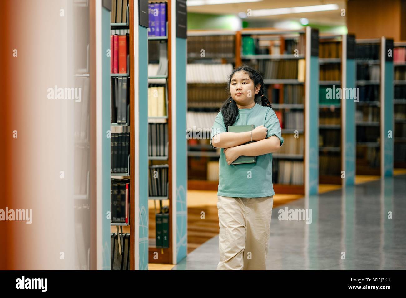 Fille asiatique portant le livre tout en marchant à l'intérieur de la bibliothèque de l'école. Posture calme communique la confiance auto-apprentissage motivation alphabétisation habitude inclusive e Banque D'Images