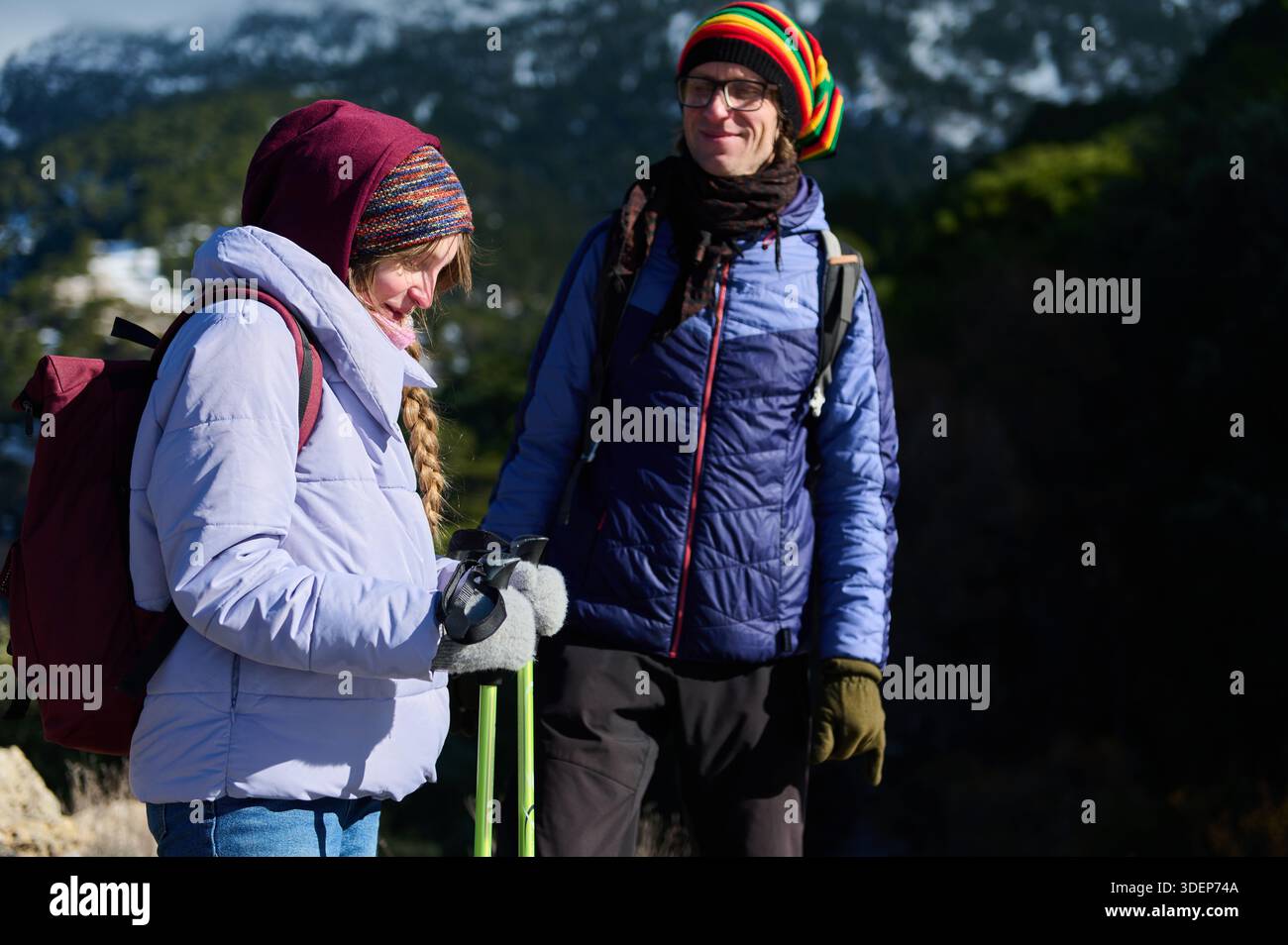 Deux randonneurs se tiennent debout sur une montagne enneigée, vêtus de vestes gonflantes, de chapeaux en tricot et de gants. Ils portent des sacs à dos et des bâtons de trekking, partageant un moment Banque D'Images