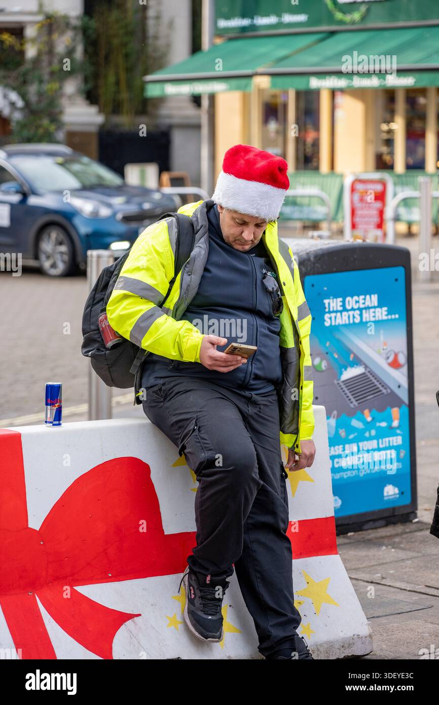 Chauffeur de bus prenant un repos portant un chapeau de Père Noël et une veste haute visibilité regardant son téléphone portable assis sur une grande boîte avec un arc rouge. Banque D'Images