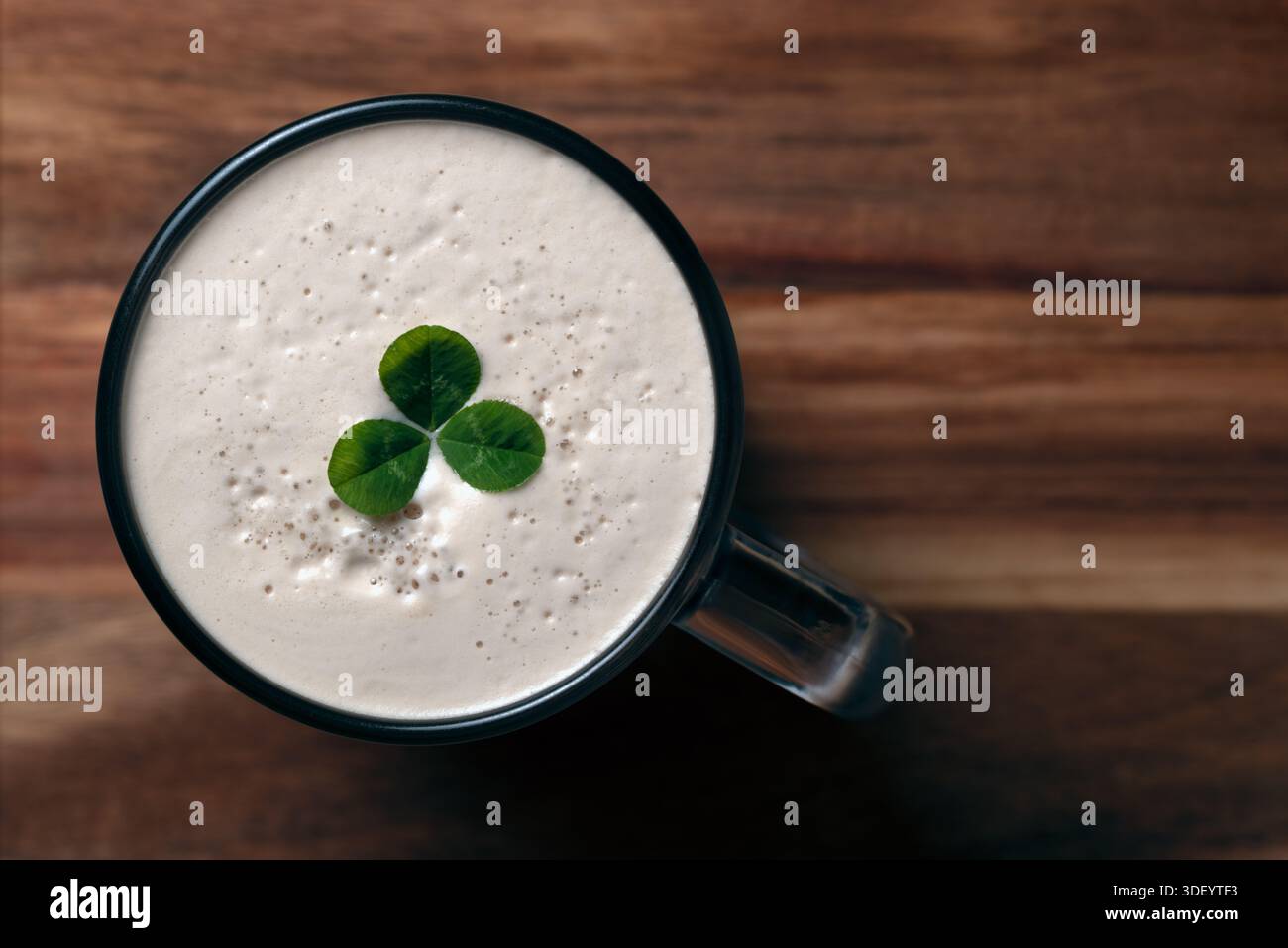 Plan à angle élevé d'une tasse de bière foncée avec une tête en mousse crémeuse décorée d'un trèfle vert à trois feuilles sur une surface en bois. La journée de Patrick Banque D'Images