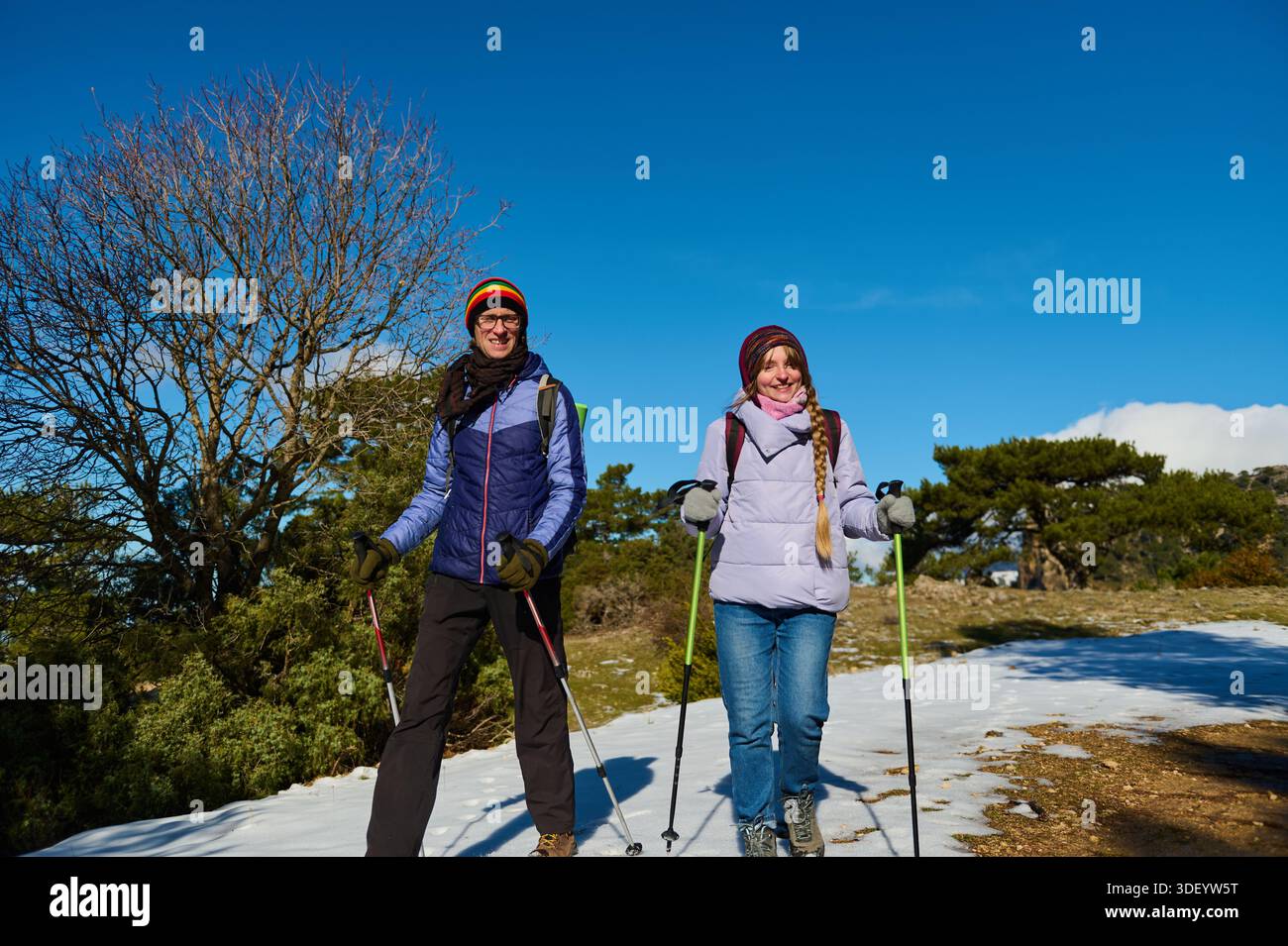 Deux randonneurs se tiennent sur un sentier enneigé sous un ciel bleu vif, souriant et habillé pour l’hiver. Ils tiennent des bâtons de trekking, prêts pour une journée d'avènement en plein air Banque D'Images