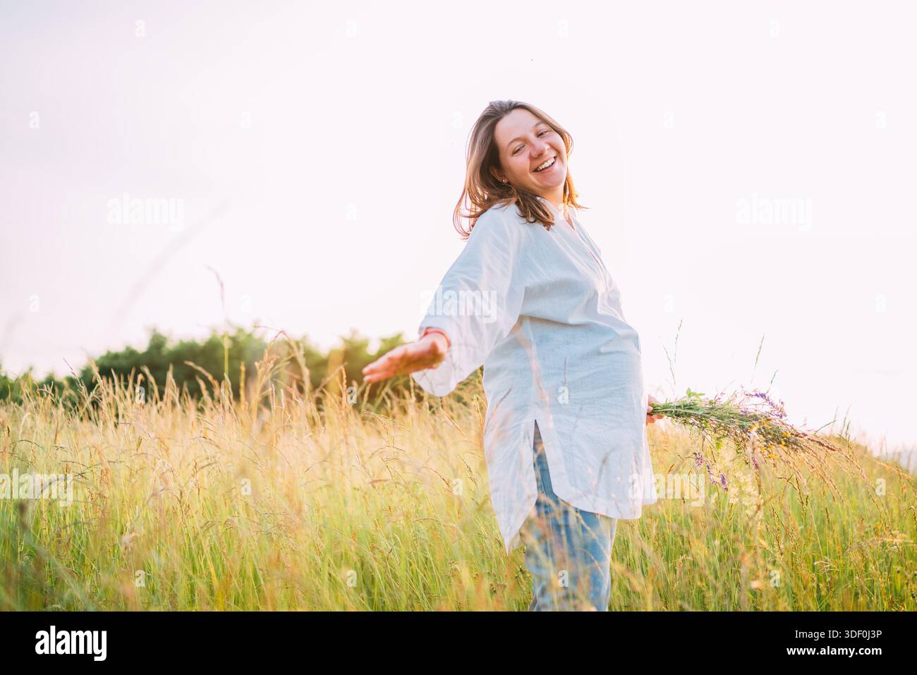 Sincèrement souriante jeune femme enceinte vêtue de vêtements d'été légers marchant près de prairie d'herbe verte haute avec bouquet de fleurs sauvages pendant le soleil du soir Banque D'Images