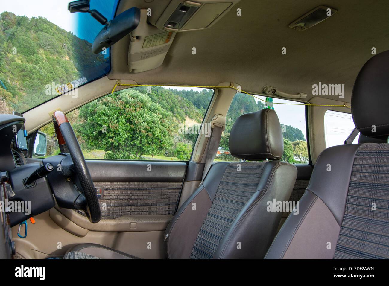 Vue intérieure d'une voiture montrant les sièges avant et le tableau de bord, avec un arrière-plan panoramique d'arbres et de collines visibles à travers le pare-brise. Banque D'Images