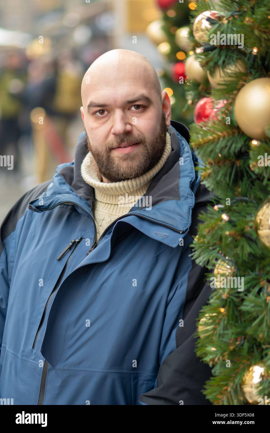 Un homme d'âge moyen barbu souriant à côté d'un arbre de Noël décoré dans la ville en hiver. Banque D'Images
