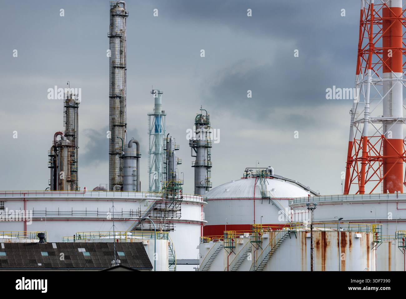Raffinerie industrielle tentaculaire avec colonnes de distillation et conduit de fumée soutenu par un treillis rouge-blanc s'élevant au-dessus des réservoirs de stockage sous un ciel couvert gris bleuâtre. Banque D'Images