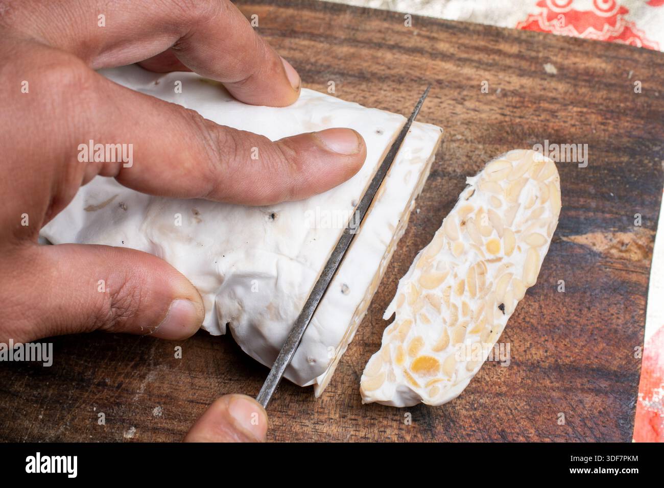 Gros plan d'une main tranchant un gâteau de soja fermenté ou du tempeh cru sur une planche de bois rustique, préparant des protéines saines à base de plantes dans une cuisine domestique. Banque D'Images