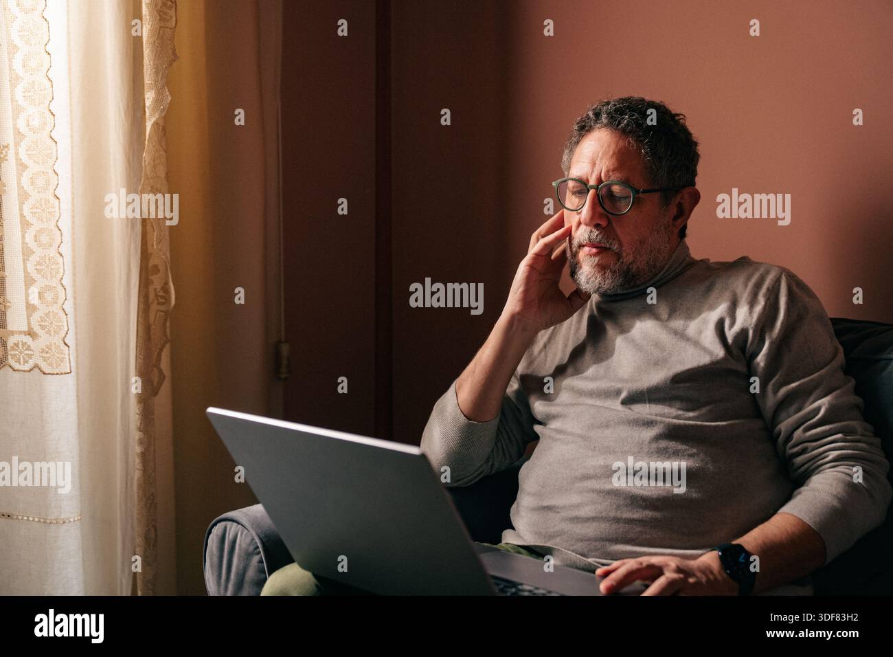 Homme senior travaillant à la maison avec un ordinateur portable près d'une fenêtre, réfléchi et stressé tout en analysant des informations à l'écran dans une lumière naturelle chaude. Banque D'Images