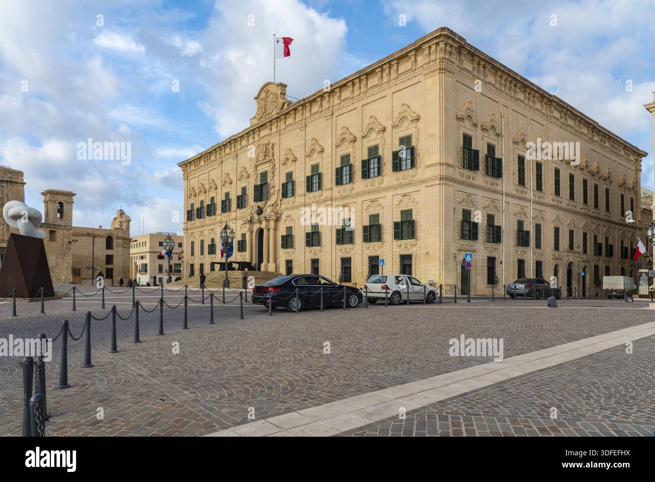 Vue de l'imposant bâtiment il-Berġa ta' Kastilja s'élève au-dessus de la place texturée tandis que les voitures sont garées, et un drapeau vole haut contre un bleu, légèrement cl Banque D'Images