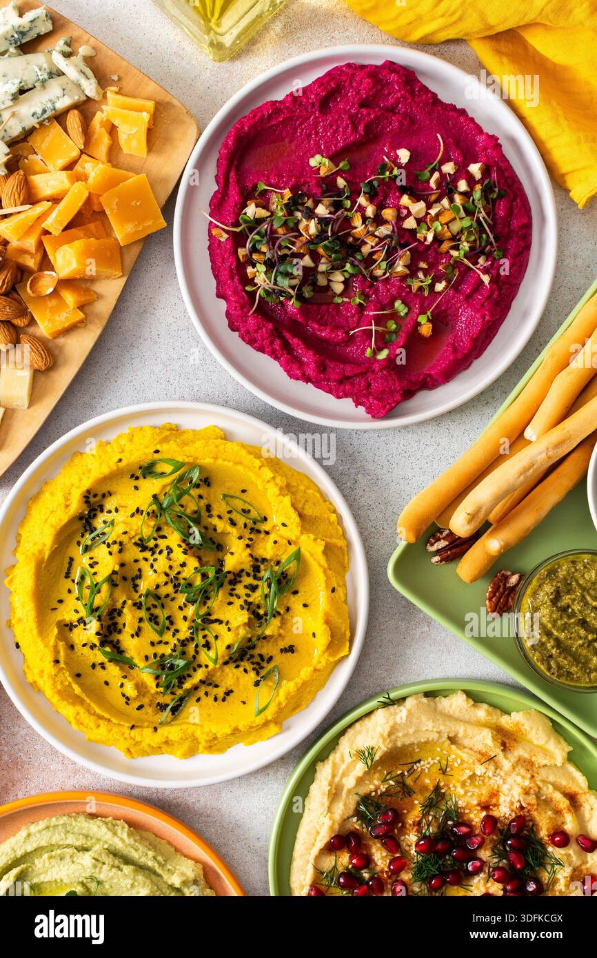 La tartinade colorée comprend des betteraves, des houmous classiques, des brocolis et du curcuma présentés sur un plateau de charcuterie avec des garnitures vibrantes, des gressins et des olives assorties, parfaits pour les rassemblements Banque D'Images