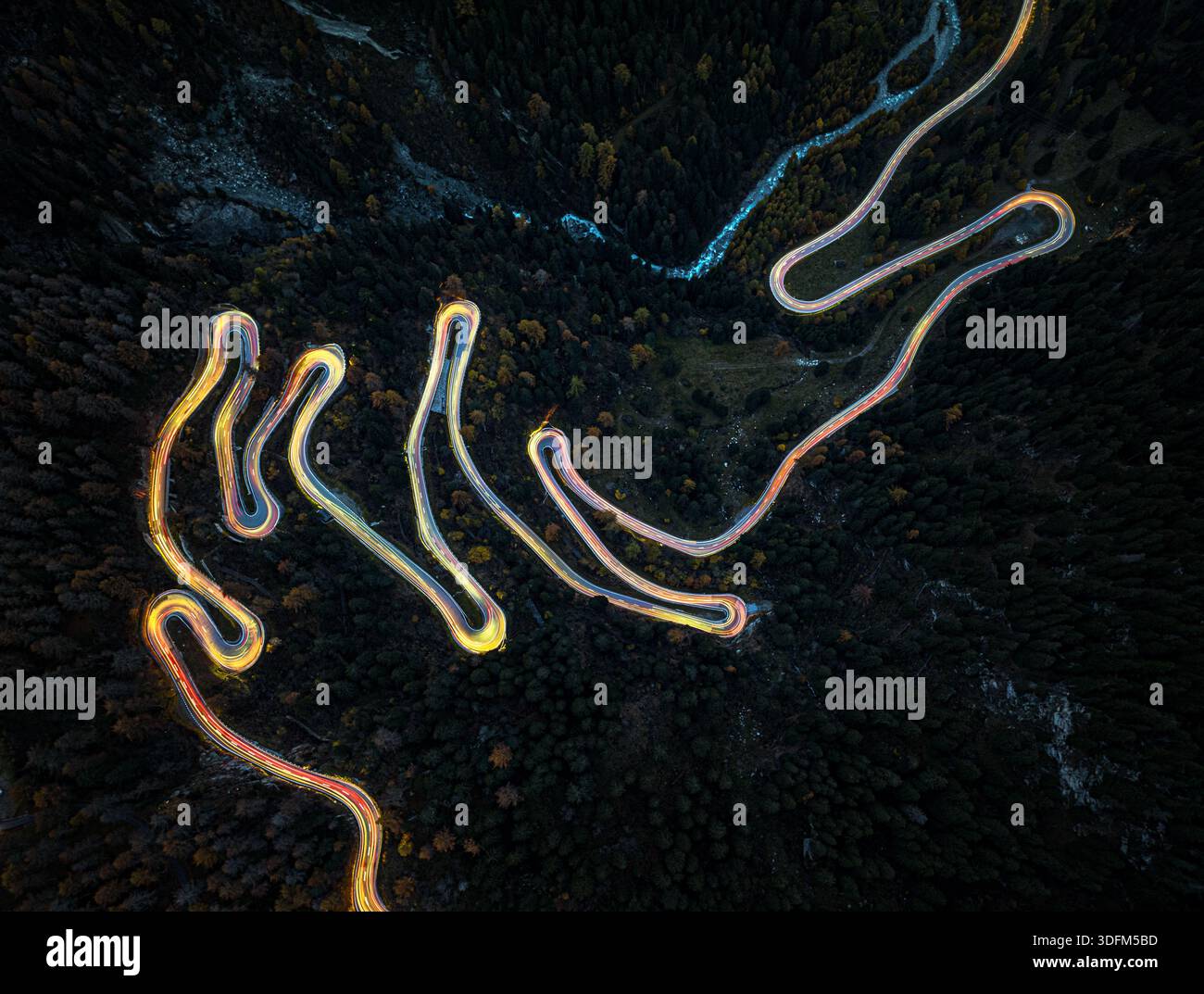 Vue aérienne d'une route sinueuse serpente à travers le terrain sombre et boisé, reflétant la lueur ambre du crépuscule sur la toile de fond enroûtante, (Malojapass) Banque D'Images