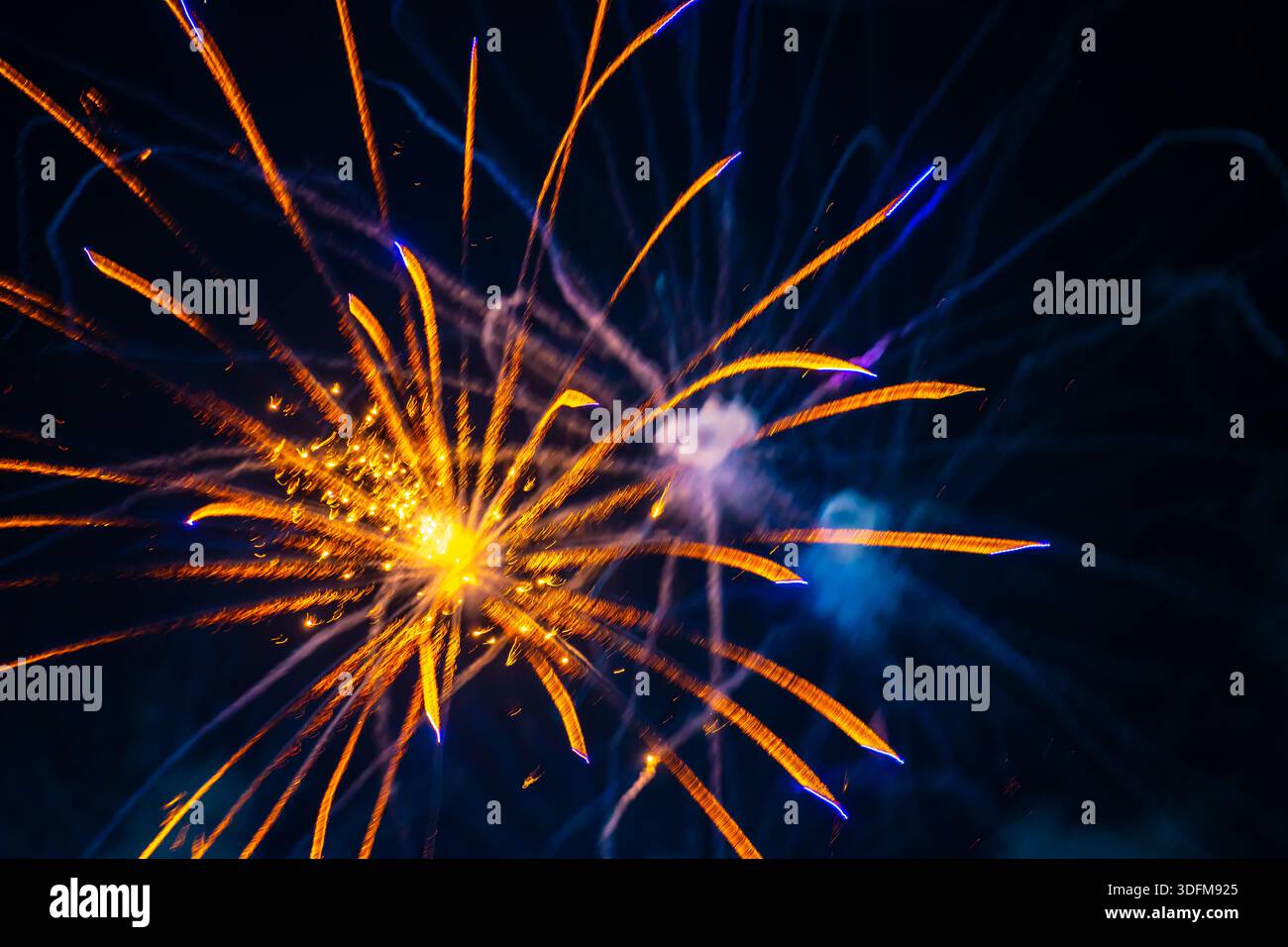 explosion vibrante de feu d'artifice dans le ciel nocturne. Des traînées dorées rayonnent vers l'extérieur à partir d'un centre lumineux. La scène comprend des notes de fumée et de lumière bleue. Banque D'Images