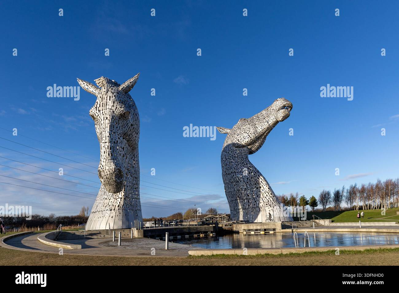 Deux imposantes sculptures de tête de cheval en acier inoxydable se dressent près d'un canal sous un ciel bleu vif, avec un paysage herbeux et des arbres en arrière-plan. Banque D'Images