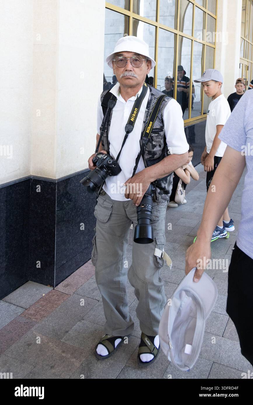 Photographe de rue asiatique âgé transportant des appareils photo en milieu urbain pendant la journée à Bichkek, Kirghizistan - 8 mai 2025 Banque D'Images
