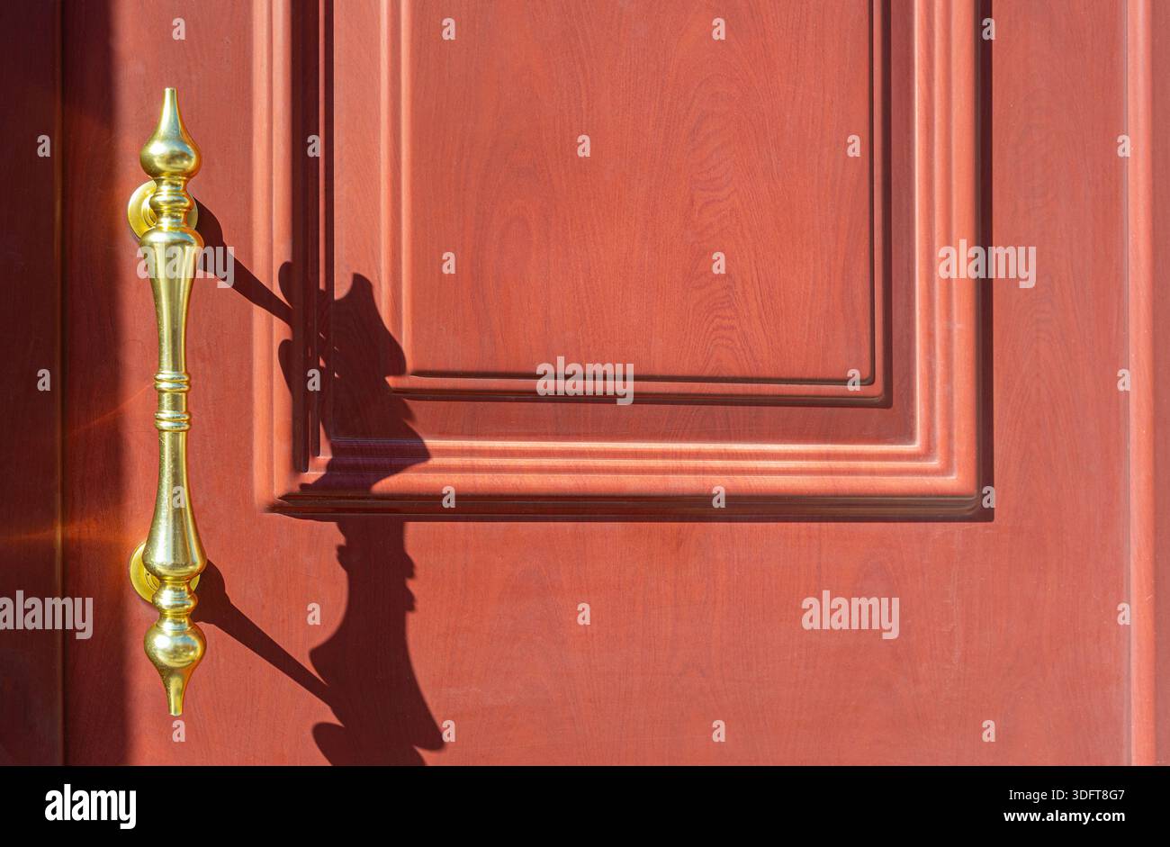 Poignée dorée projetant une longue ombre sur la surface de la porte en bois rouge, créant un motif décoratif Banque D'Images