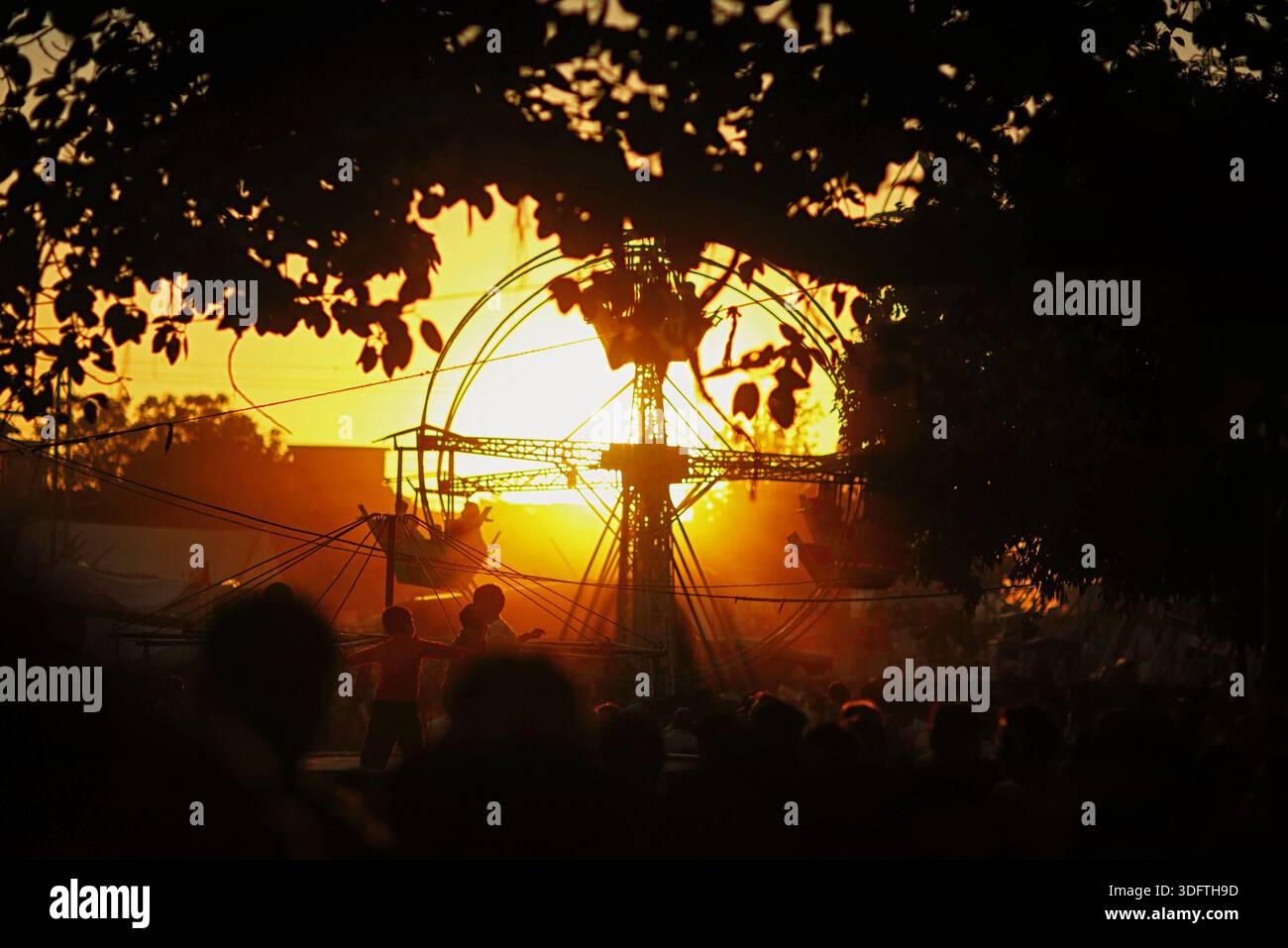 Lahore, Pakistan - 24 décembre 2025 : vue du soleil se couchant derrière la grande roue silhouette, projetant une lueur orange chaude sur la foule à la fai Banque D'Images