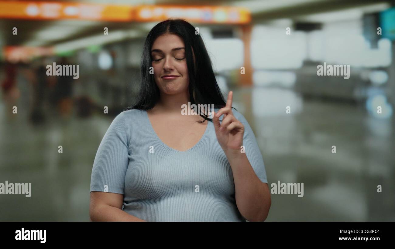 Une femme fait des gestes niant tout en se pointant vers elle-même dans un terminal d'aéroport occupé, exprimant sa curiosité et véhiculant une communication non verbale dans un Banque D'Images