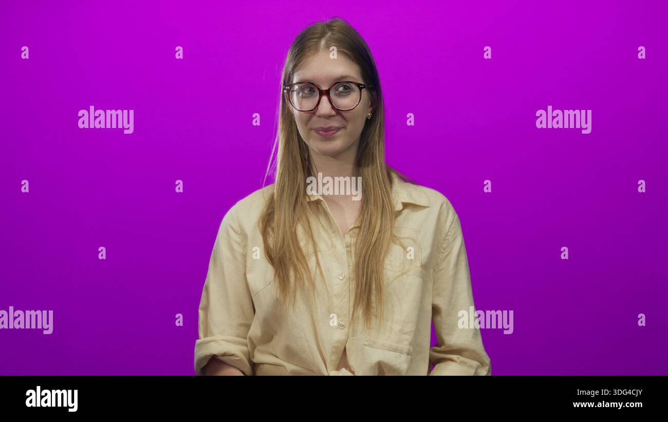 Jeune femme blonde portant une chemise beige tenant un cœur rouge avec les deux mains et souriant dans un studio violet vibrant ; affection. Banque D'Images