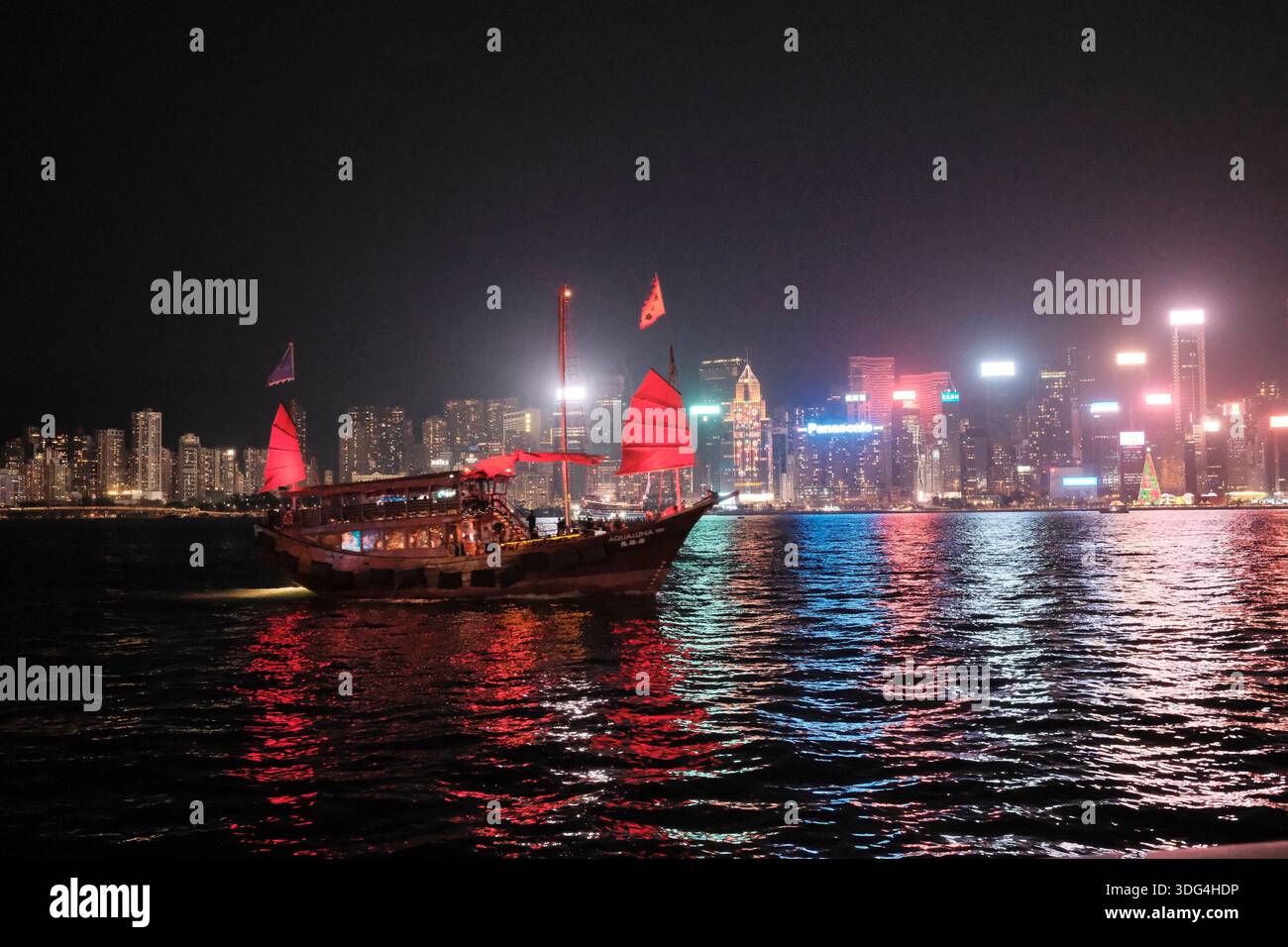 Une jonque chinoise traditionnelle traverse le port Victoria la nuit, avec les gratte-ciel illuminés de Hong Kong qui se reflètent dans l'eau derrière Banque D'Images