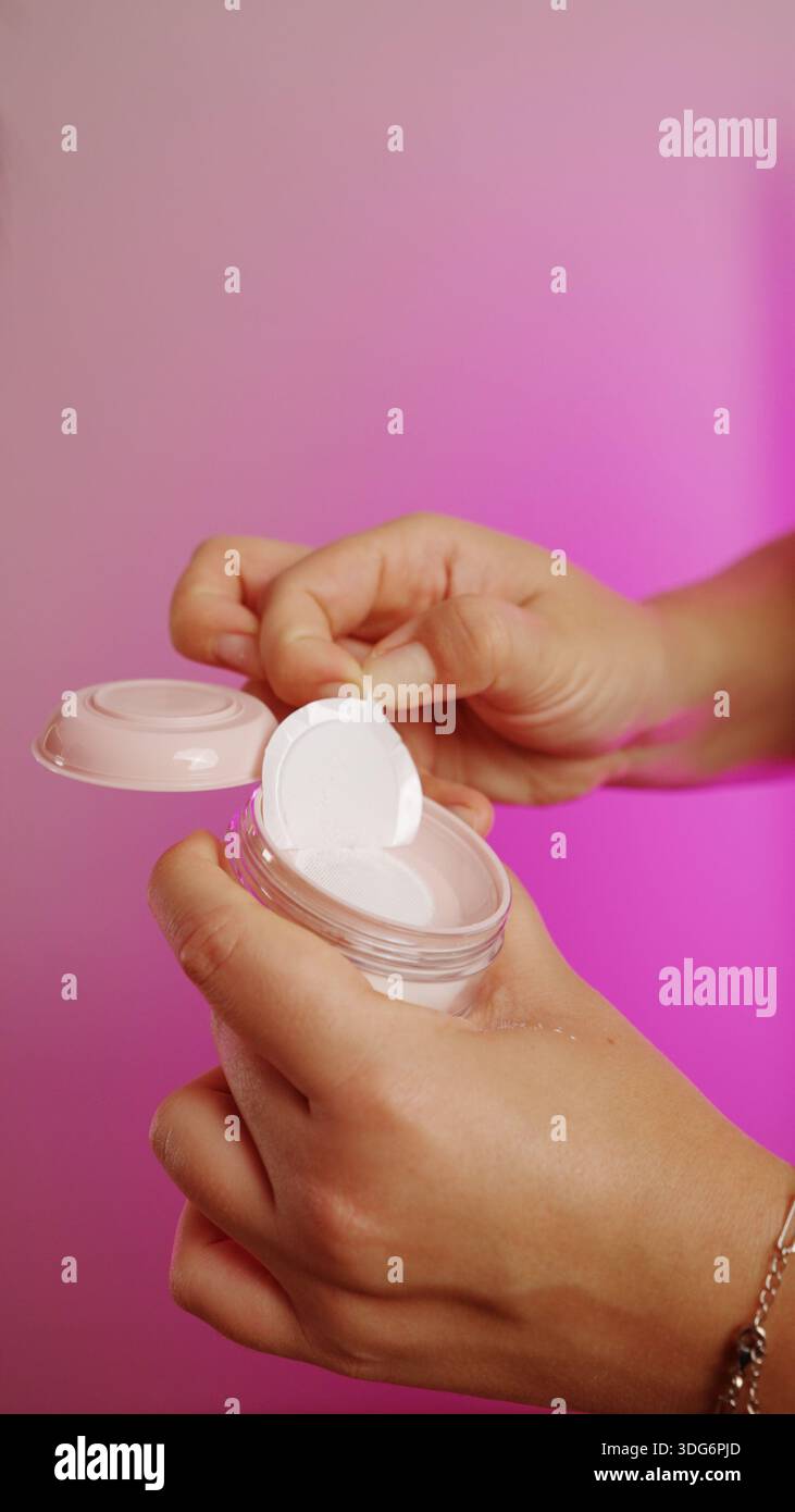 Gros plan des mains ouvrant ou tenant un petit récipient cosmétique sur un fond rose vif. Banque D'Images