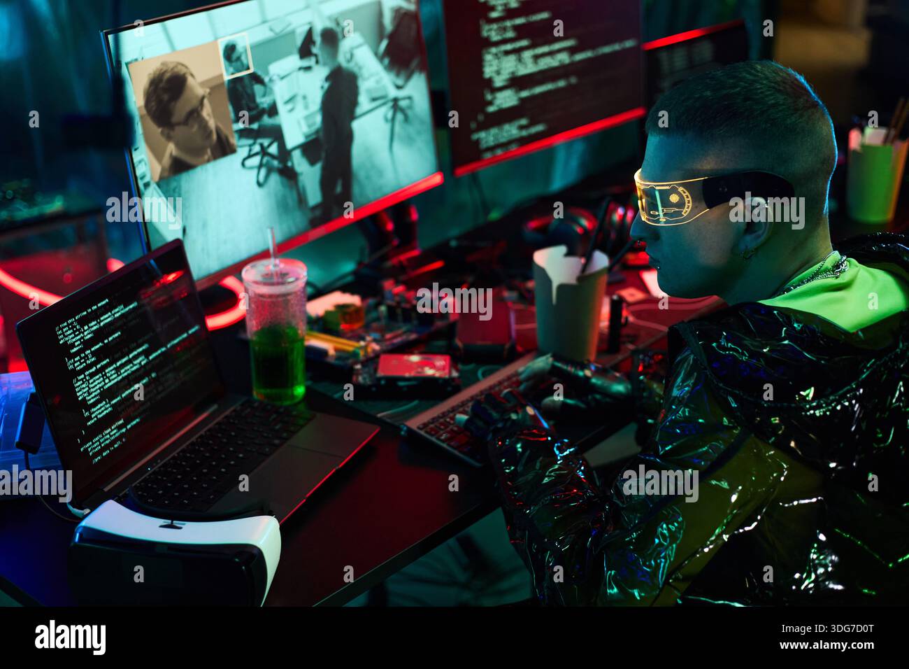 Jeune homme caucasien adulte portant des lunettes futuristes assis au bureau encombré tapant sur le clavier, plusieurs moniteurs affichant le code et les séquences de surveillance, se livrant à une activité de piratage Banque D'Images