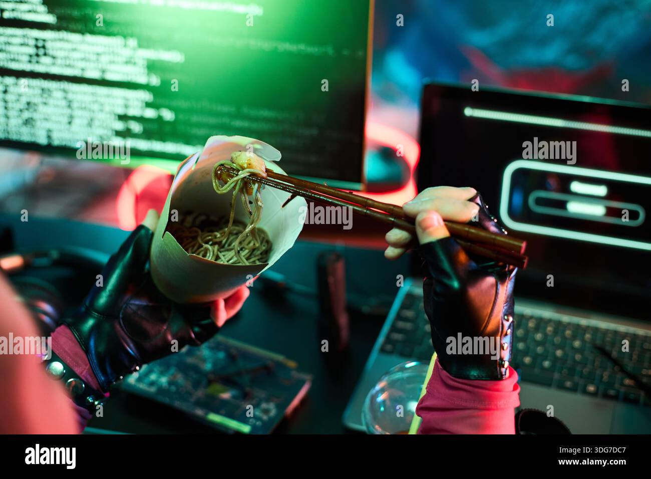 Jeune femme caucasienne portant des gants noirs, assise au bureau mangeant des nouilles avec des baguettes, tout en travaillant sur des écrans d'ordinateur portable et d'ordinateur, affichant le code de piratage malveillant dans une pièce sombre Banque D'Images