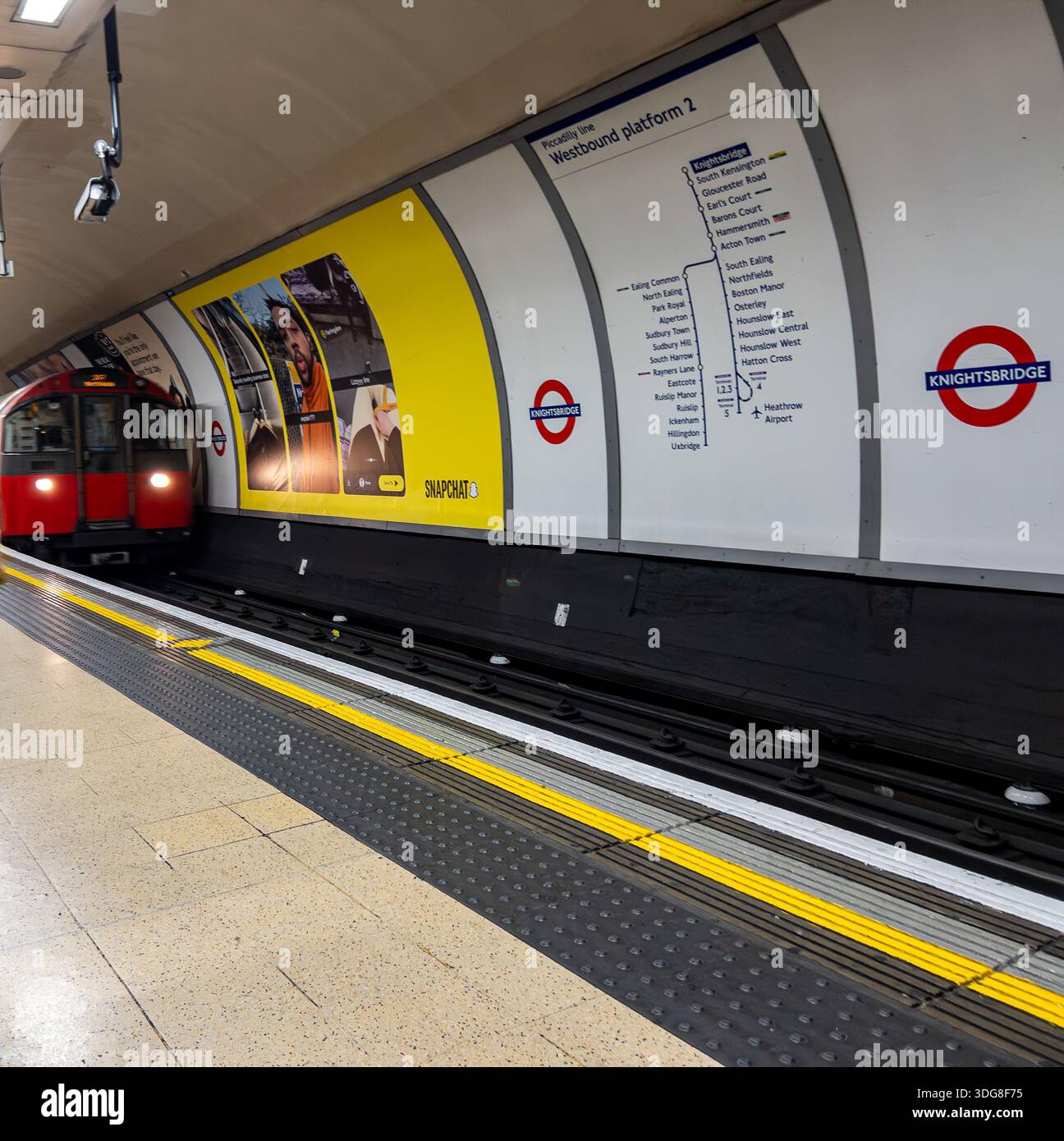 Train de métro londonien arrivant à la station Knightsbridge sur la ligne Piccadilly avec signalisation du tunnel et publicité visible sur le mur du quai Banque D'Images