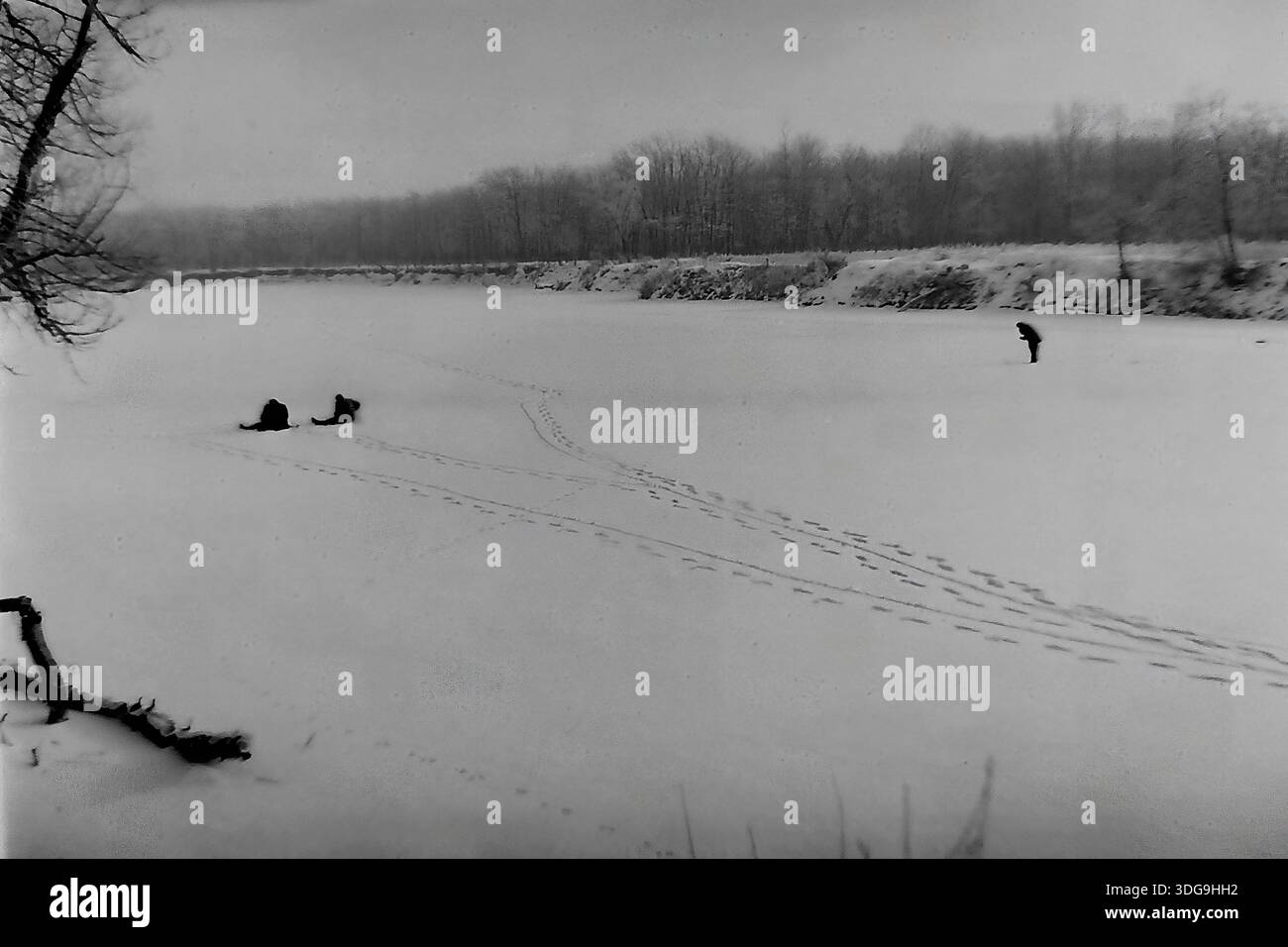 Slavyansk, région de Donetsk, Ukraine, URSS. Archive photo en noir et blanc des années 1960 capturant un vaste paysage hivernal sur une rivière gelée. La vaste surface blanche est parsemée de trois petites figures de pêcheurs de glace : deux sont assis près de leurs trous, tandis que l'une se tient plus loin. Des empreintes de pas distinctes sillonnent la neige. Au premier plan, un accroc sombre dépasse du banc de neige. Une ligne de forêt dense crée une toile de fond sous le ciel gris. Cette image atmosphérique illustre la solitude tranquille de la pêche hivernale dans la paisible région du Donbass. Banque D'Images