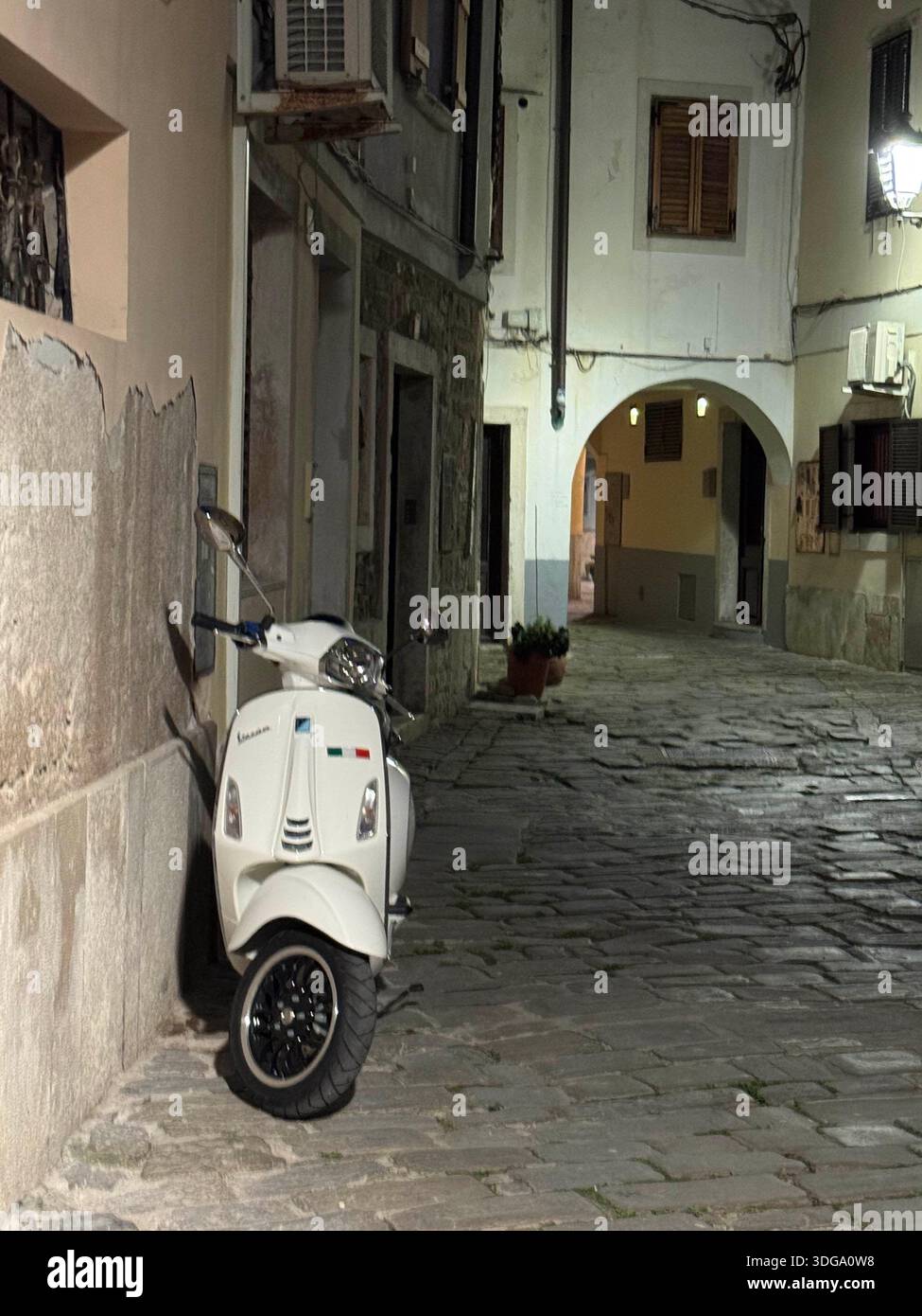 Vue nocturne atmosphérique d'une rue pavée étroite à Piran, Slovénie, avec un scooter Vespa classique Banque D'Images