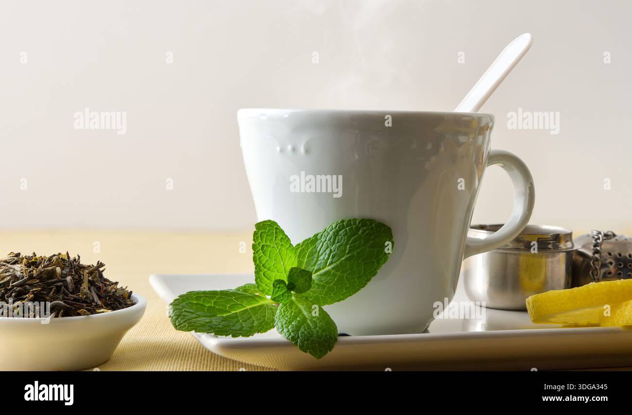 Macro détail d'une tasse de thé sur une soucoupe en céramique à la menthe et au citron sur une table avec une nappe beige, des feuilles de thé, et un infuseur, isolé sur un b blanc Banque D'Images