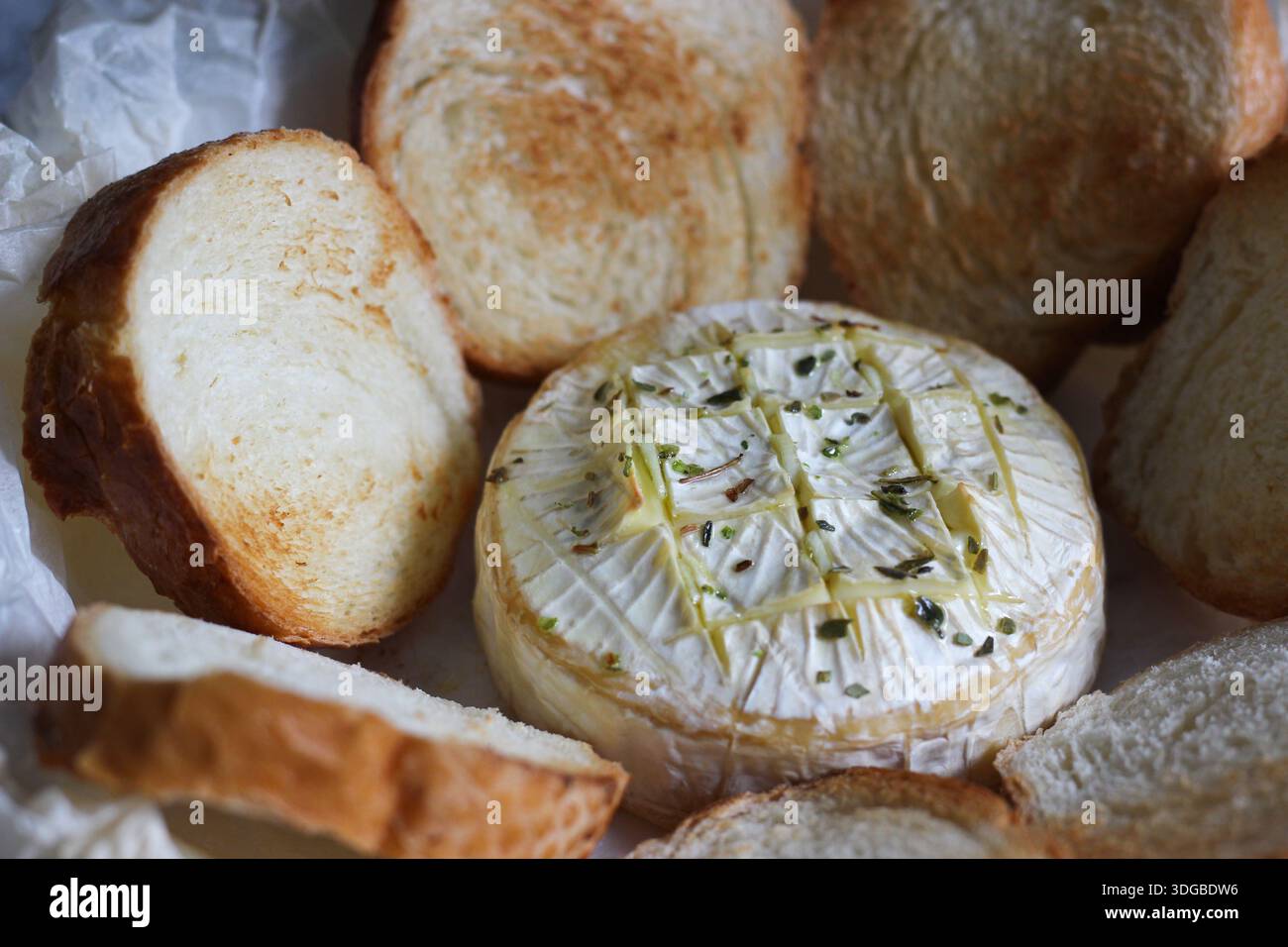 Camembert cuit au four aux herbes et à l'huile d'olive, servi avec des tranches de pain grillées sur du papier sulfurisé. Apéritif maison confortable photographié en naturel Banque D'Images