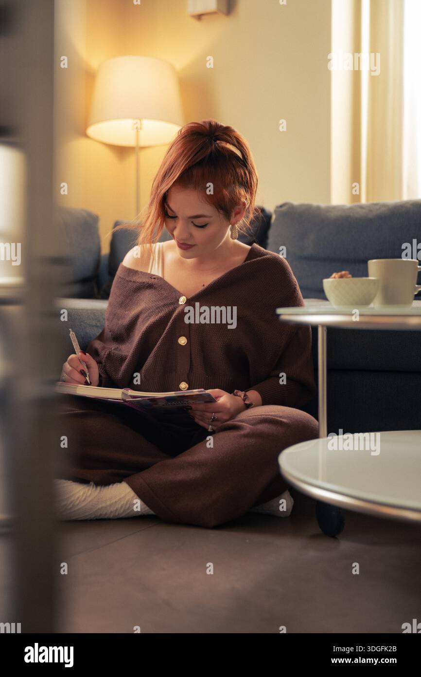 Une femme calme est assise sur le sol à la maison tout en écrivant dans son journal personnel, profitant d'un moment calme de pleine conscience, de réflexion personnelle et de vie lente Banque D'Images
