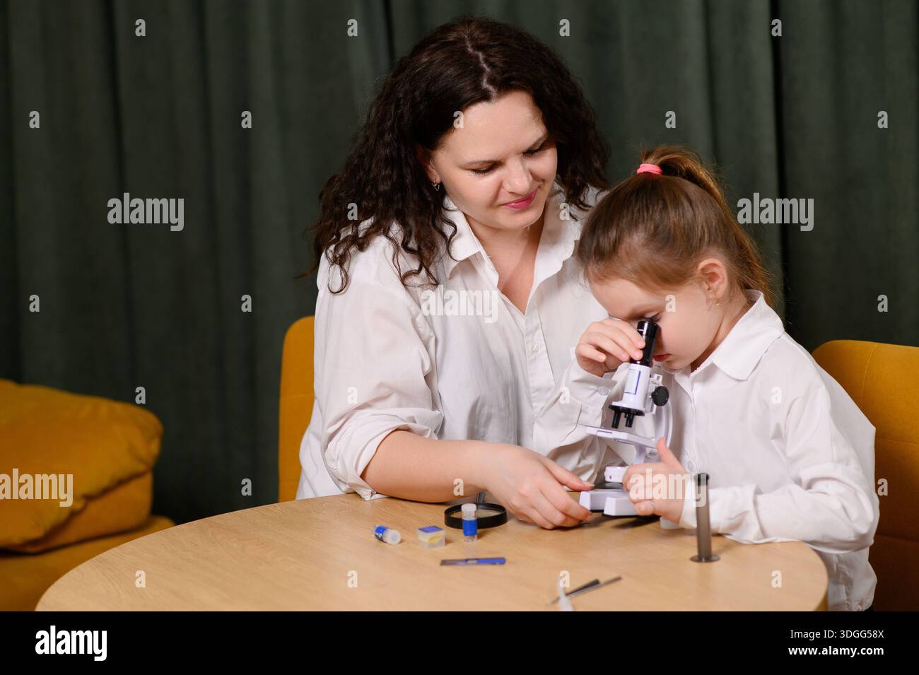 Maman et fille examinent les spécimens au microscope tout en étant assis dans le salon. La femme aide la fille à mener des expériences scientifiques pratiques en utilisant micro Banque D'Images