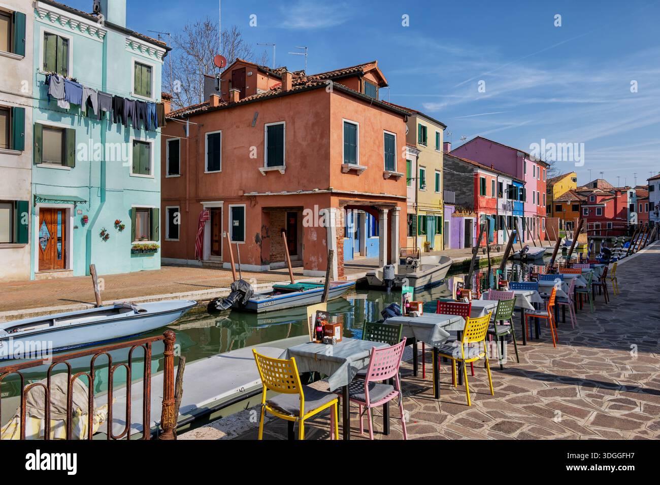 Île de Burano dans la lagune vénitienne, Italie. Tables de restaurant prêtes pour les clients sur la promenade au canal avec vue sur les maisons traditionnelles avec fa peint Banque D'Images