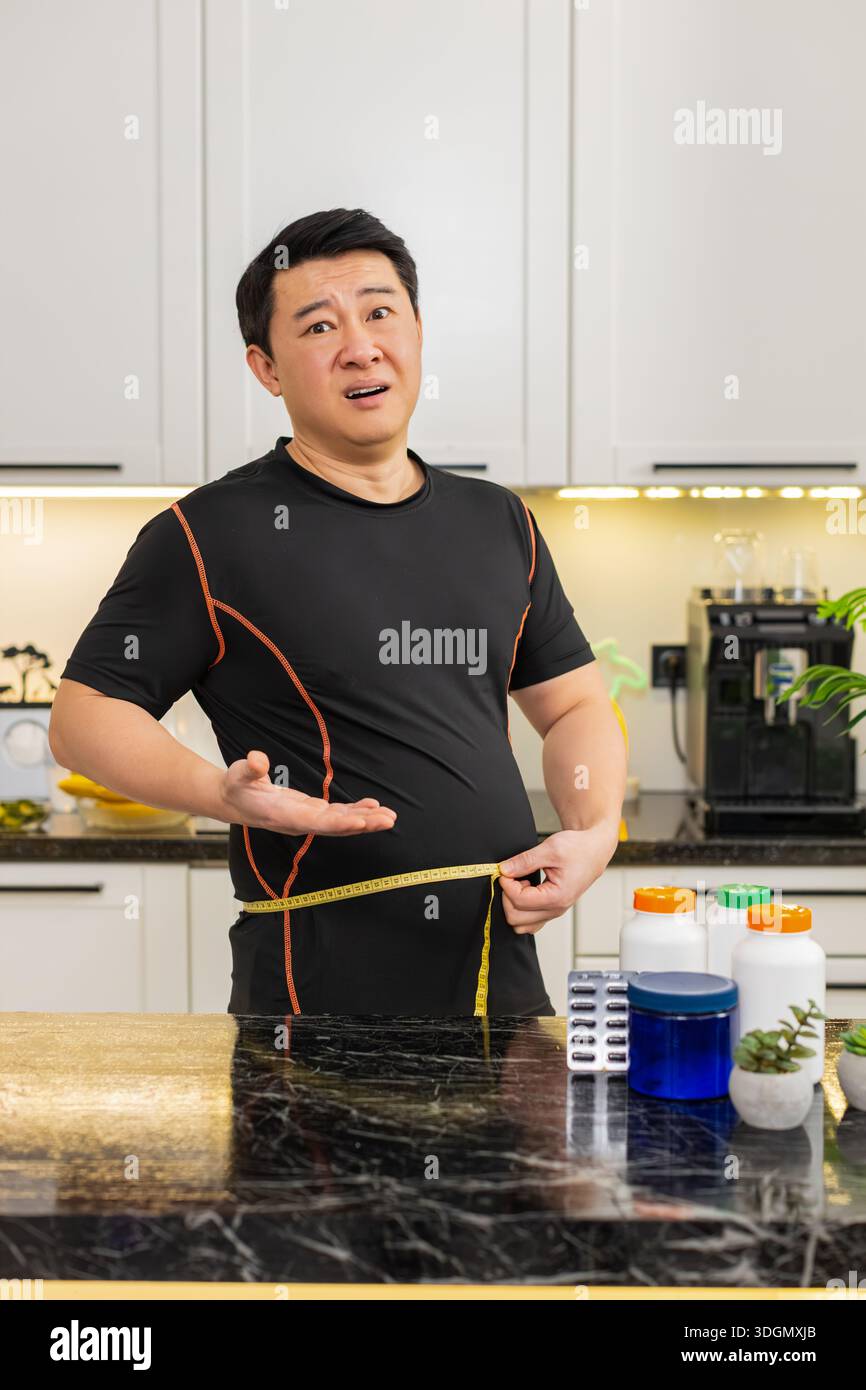 L'homme asiatique en surpoids dans la cuisine à la maison mesure la taille avec du ruban adhésif et semble frustré par le gros ventre après le gain de poids. Les capsules de régime sur la table suggèrent la recherche pour le changement de routine de santé de soutien amincissant. Banque D'Images