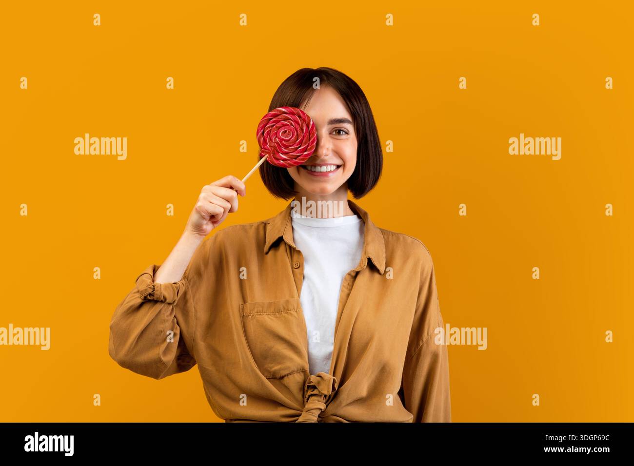 Concept de dent douce. Bonne jeune femme tenant un bonbon sur bâton, couvrant un oeil avec un lollipop, fond jaune studio Banque D'Images