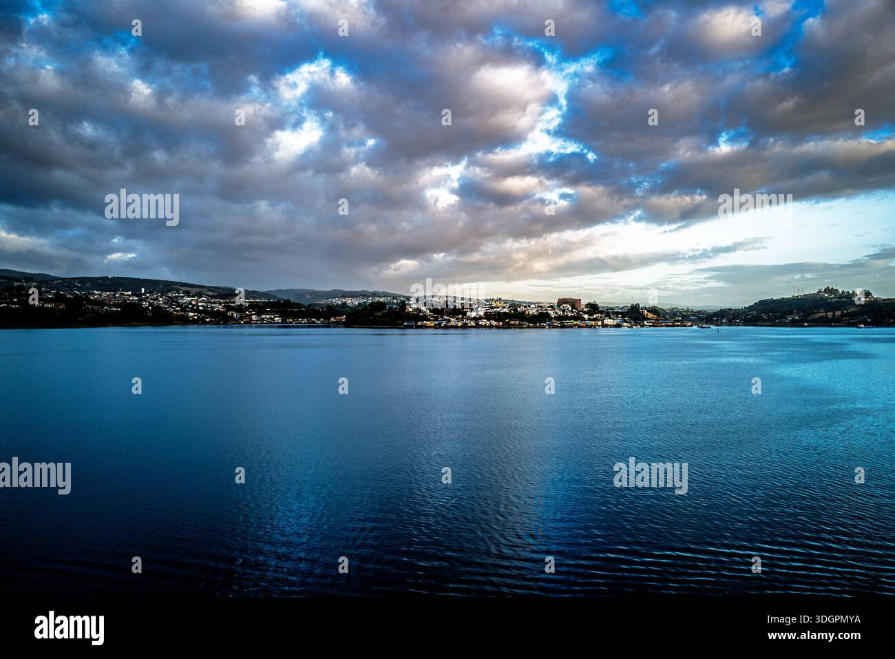 Une vaste étendue d'eau bleu profond s'étend vers une ville lointaine nichée sur des collines ondulantes sous un ciel spectaculaire rempli de nuages sombres et texturés Banque D'Images