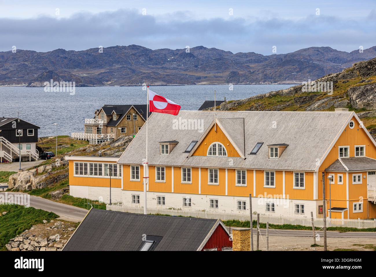 vue du paysage côtier du vieux nuuk et le drapeau du groenland sur un haut mât de drapeau devant un grand bâtiment jaune avec des montagnes en arrière-plan Banque D'Images