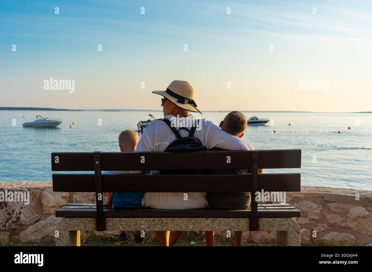 Vue arrière de la mère en chapeau de paille avec deux fils assis sur le banc regardant la mer avec des bateaux au coucher du soleil. Concept de vacances en famille, voyage méditerranéen, Banque D'Images
