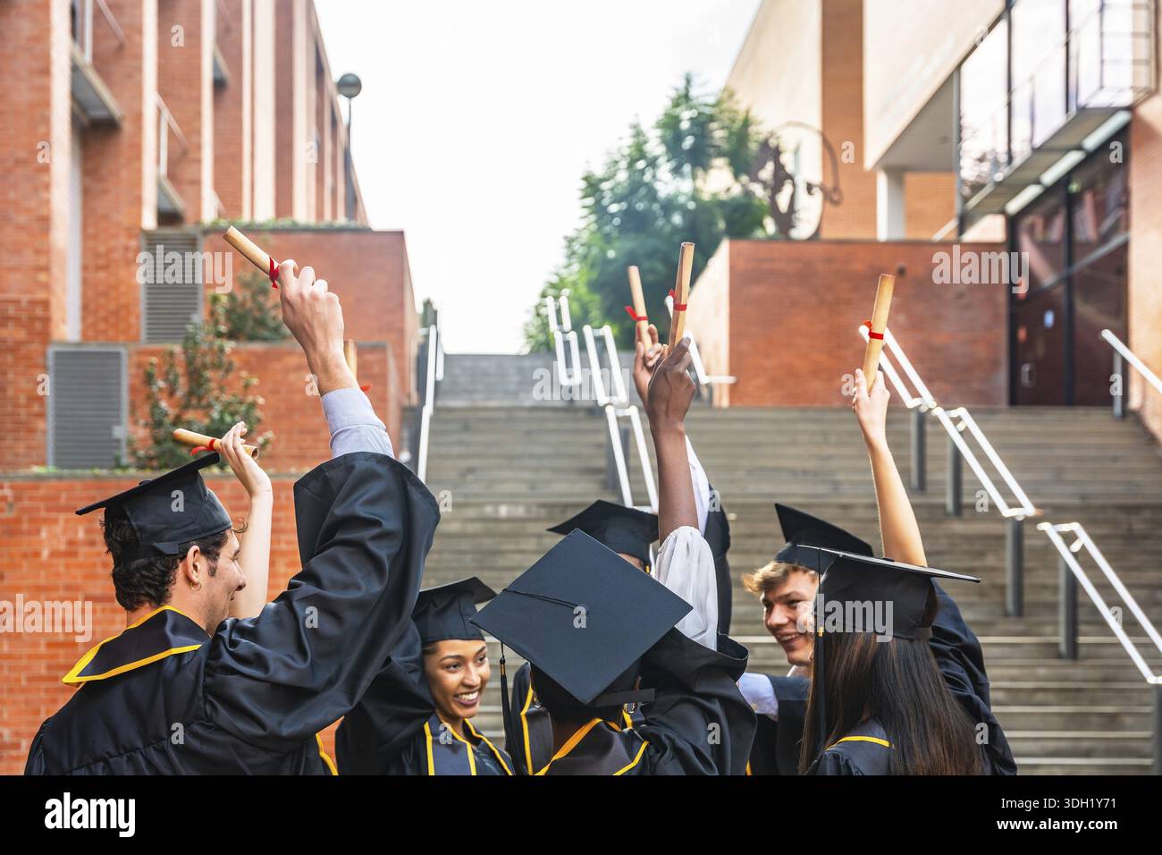 Groupe d'étudiants diplômés divers portant des casquettes et des robes académiques, élevant heureusement leurs rouleaux de diplôme dans la célébration sur un campus universitaire afte Banque D'Images