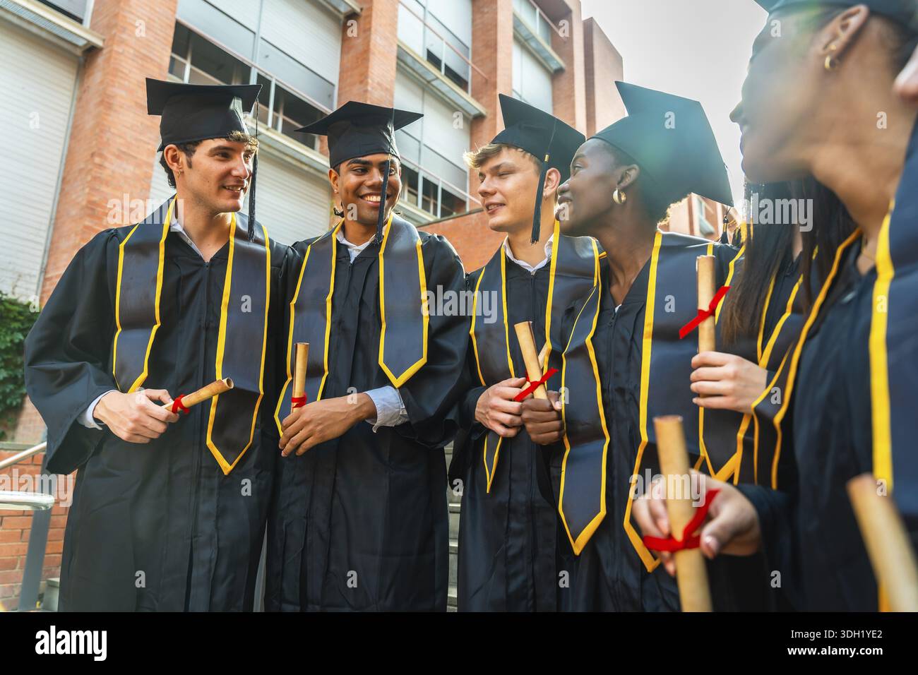Groupe d'étudiants diplômés divers portant des robes académiques et des casquettes, souriant tout en tenant leurs diplômes et célébrant à l'extérieur d'un bâtiment universitaire Banque D'Images