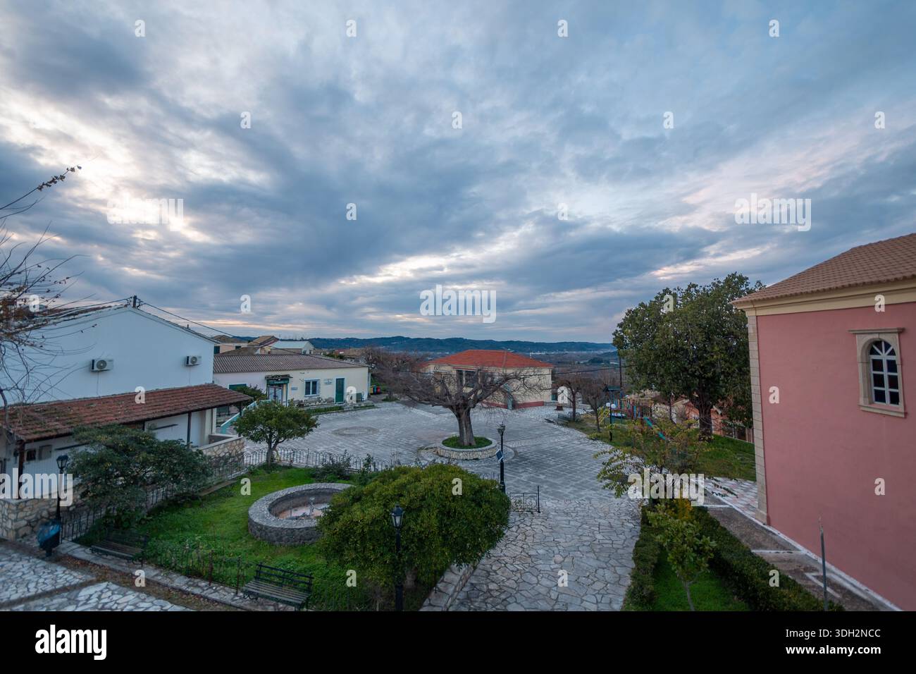 Vue grand angle du centre du village d'Agioi Douloi à Corfou, en Grèce, montrant l'église rose, les maisons traditionnelles blanches, le trottoir de pierre, et les arbres d'hiver un Banque D'Images