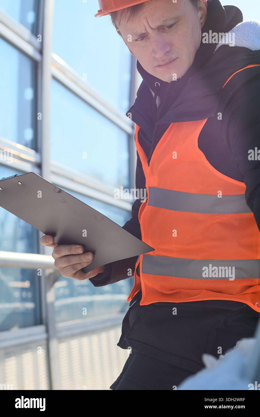 Travailleur en gilet orange et casque lit les notes sur le bloc-notes sur le lieu de construction Banque D'Images
