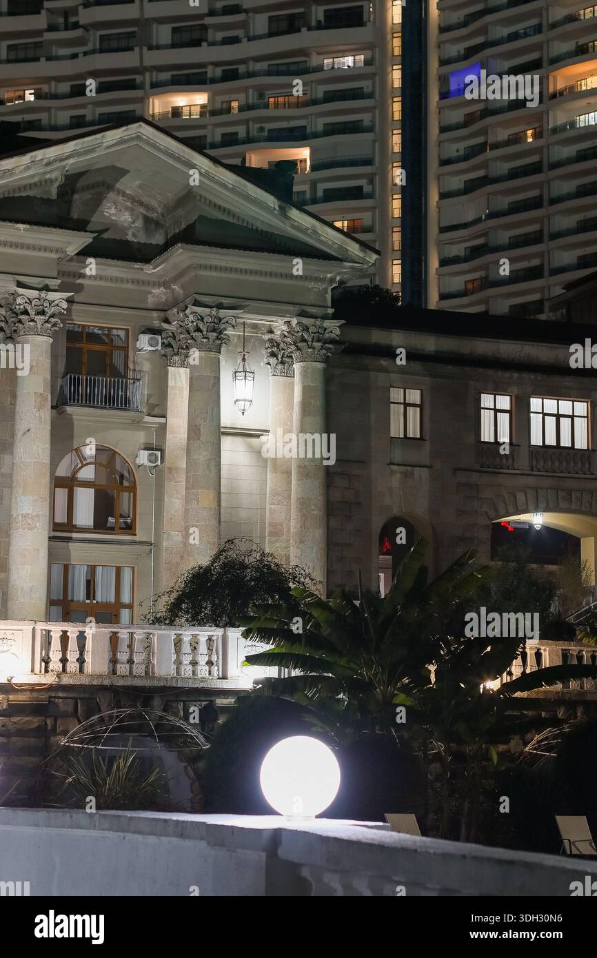 Façade néoclassique illuminée avec de hautes colonnes corinthiennes et fronton la nuit, immeubles d'appartements modernes s'élevant derrière, contraste de l'architecture Banque D'Images