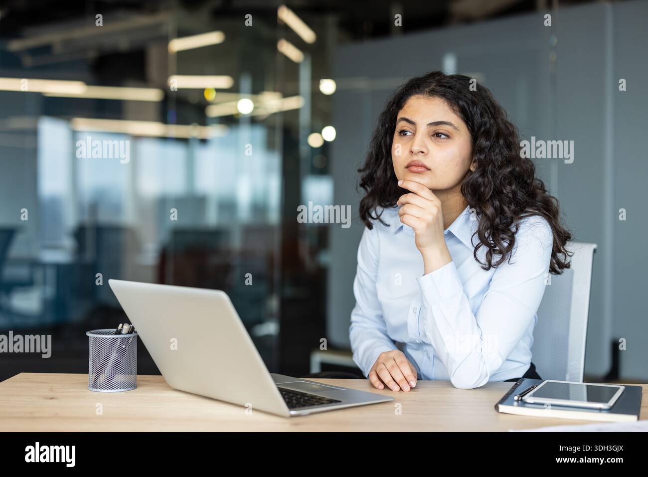 Jeune femme d'affaires aux cheveux bouclés foncés assis à un bureau avec un ordinateur portable, profondément imprégnée de pensée et concentrée sur ses ambitions professionnelles et son succès Banque D'Images