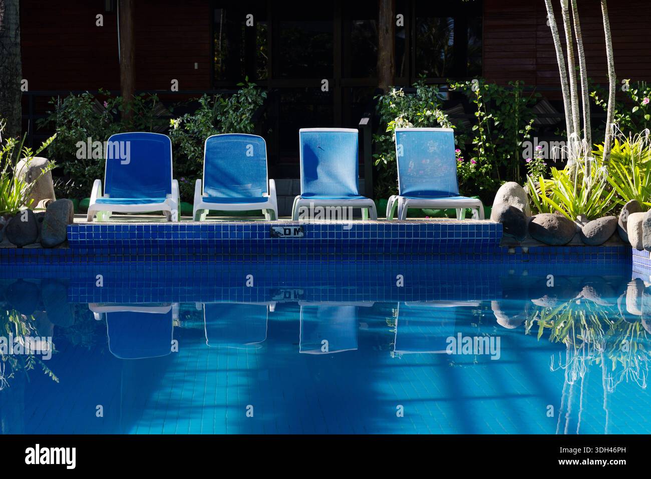 Des chaises longues bleues vides bordent une piscine tranquille d'un complexe tropical des Fidji, reflétées dans l'eau plate sous la lumière vive du matin. Banque D'Images