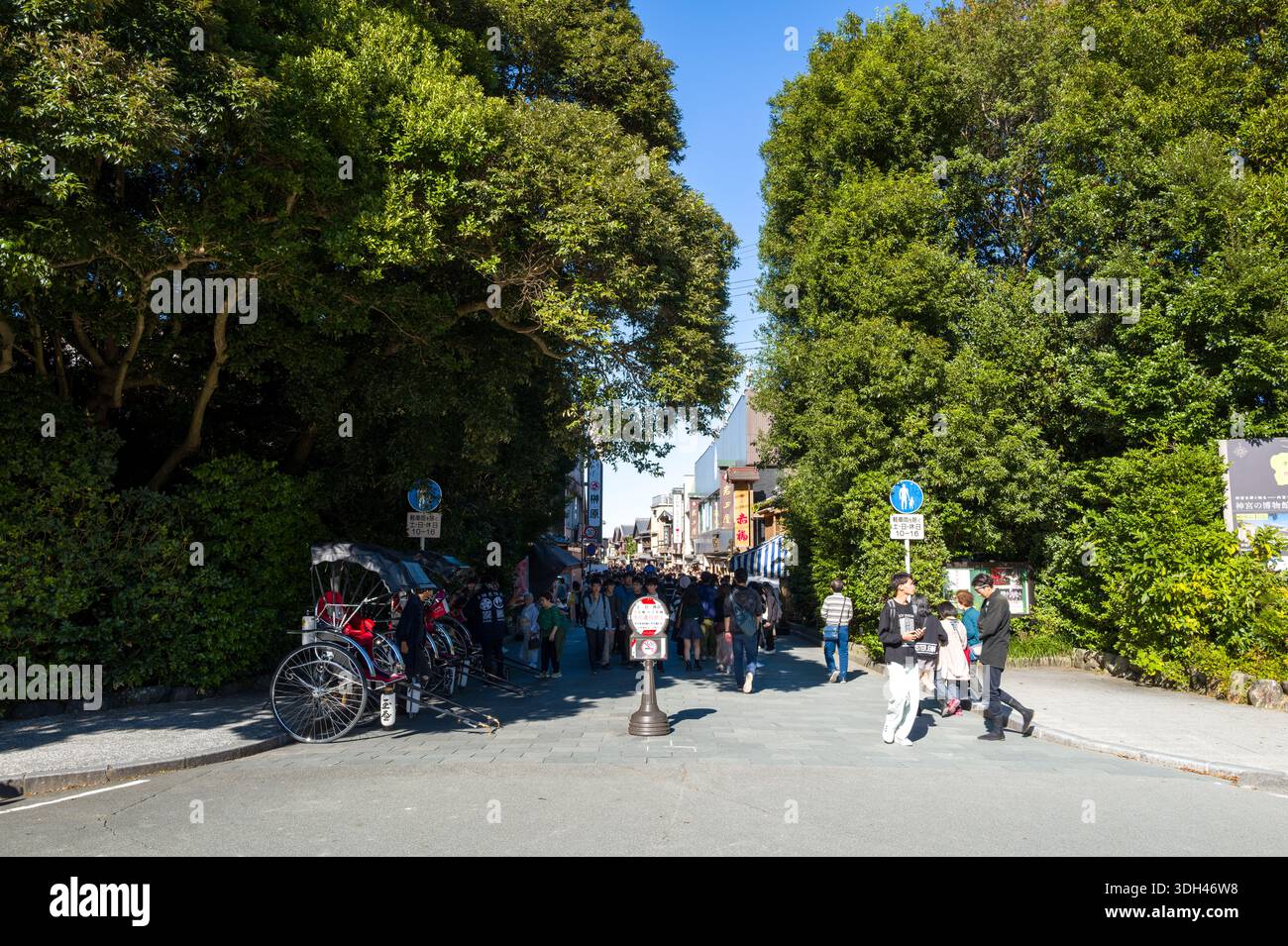 ISE, Japon - 29 octobre 2023 : des groupes de personnes marchent dans le district d'Okage Yokocho, avec des pousse-pousse alignés sur le côté gauche, de grands arbres au-dessus et des panneaux visibles sous un ciel bleu clair. Banque D'Images